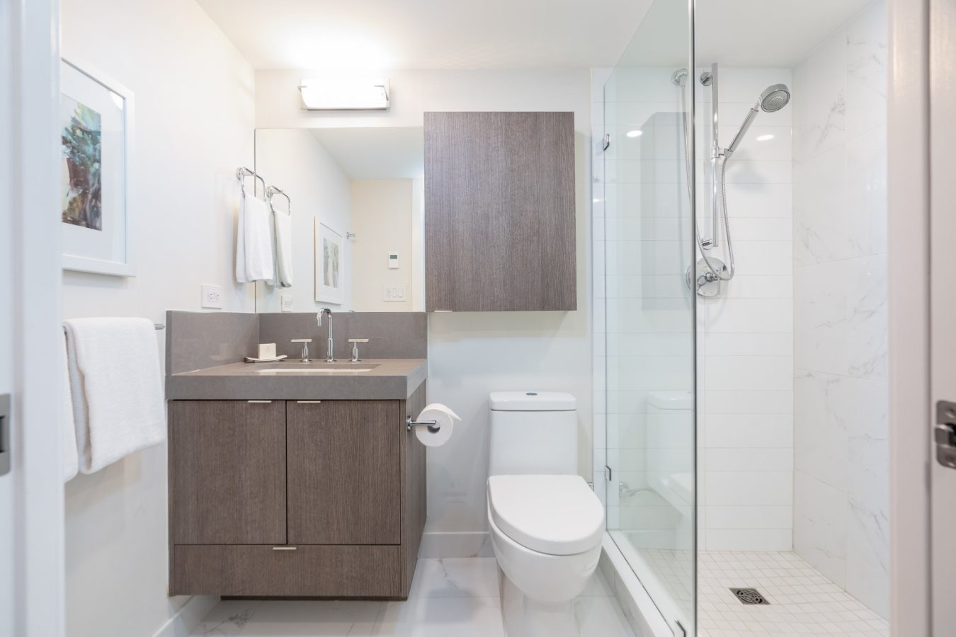 A modern bathroom with a wooden vanity, rectangular sink, wall-mounted mirror cabinet, white toilet, glass shower enclosure, white tile walls, and towel racks with white towels.