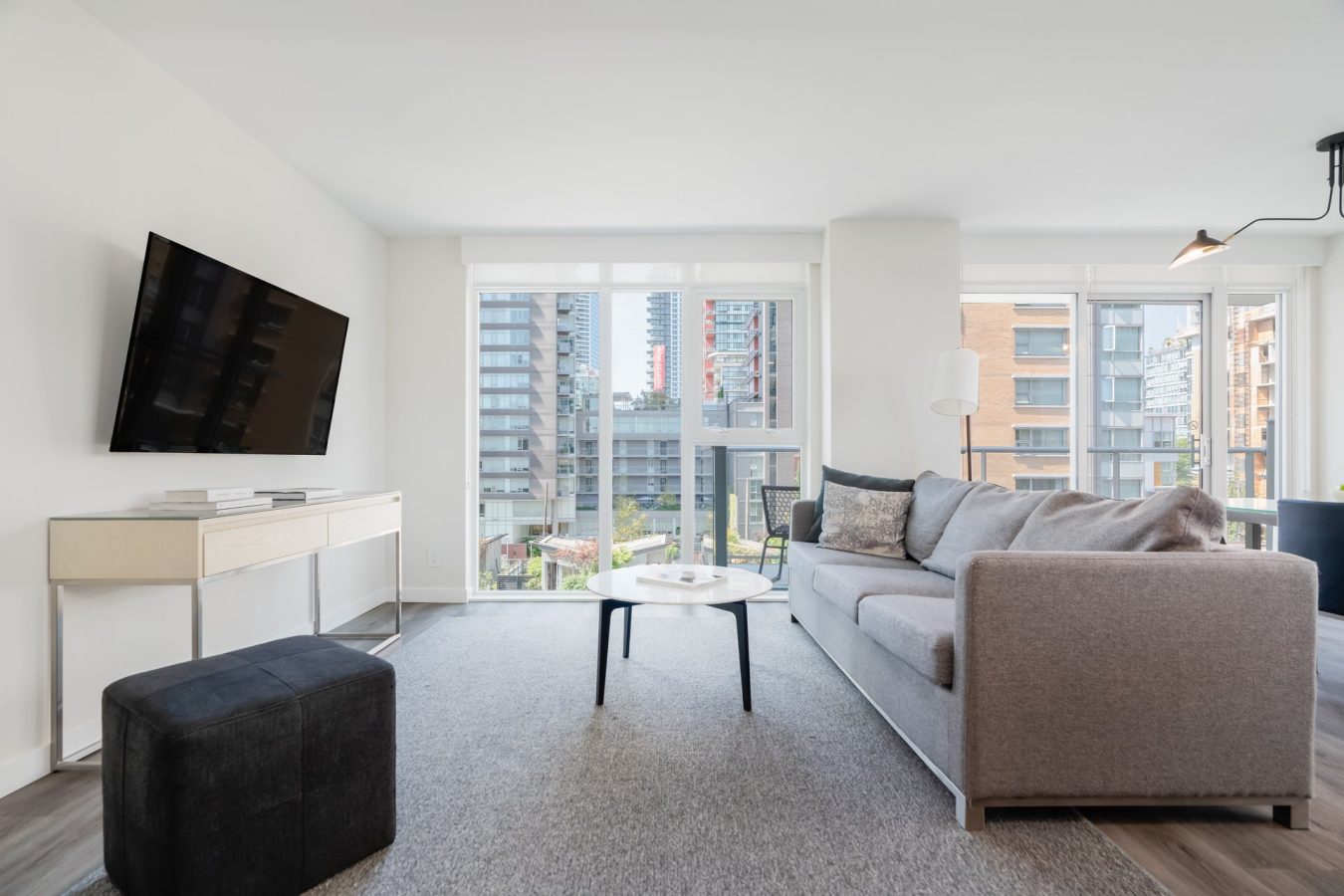 Modern living room with gray sofa, coffee table, wall-mounted TV, ottoman, and large windows offering a view of tall city buildings and letting in natural light.