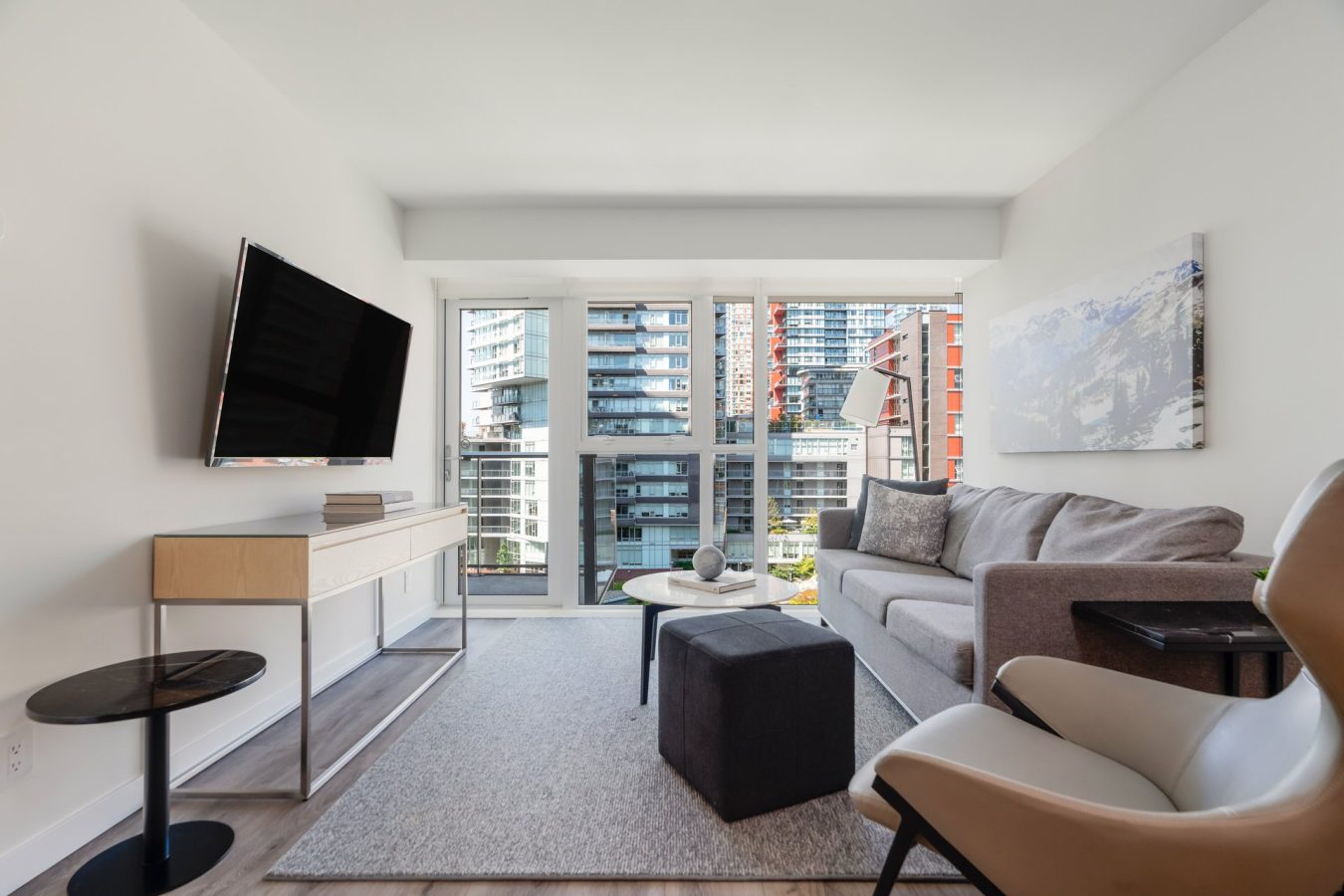 Modern living room with a gray sofa, armchair, wall-mounted TV, and a large window offering a view of tall city buildings. Neutral decor with a light rug, small tables, and wall art.