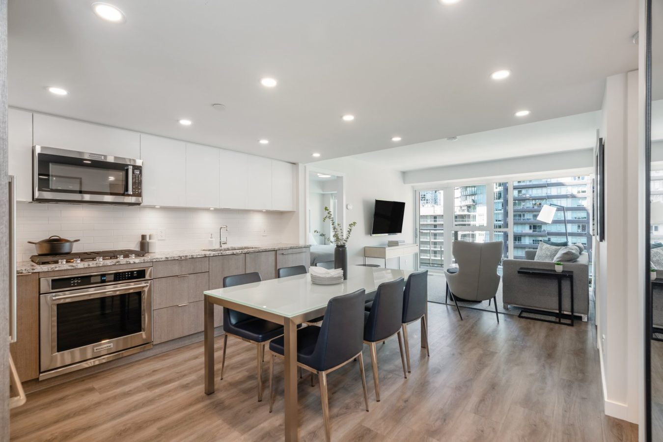 Modern open-concept kitchen and living room with wood floors, stainless steel appliances, a dining table with six chairs, large windows, and a view of nearby buildings. The space is bright with recessed lighting.
