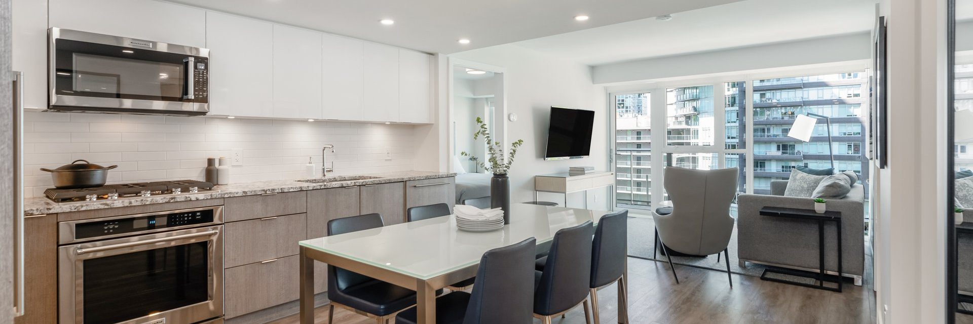 Modern open-concept kitchen and living room with light wood cabinets, stainless steel appliances, dining table with six chairs, and large windows showing city buildings outside. Neutral colors and contemporary decor throughout.