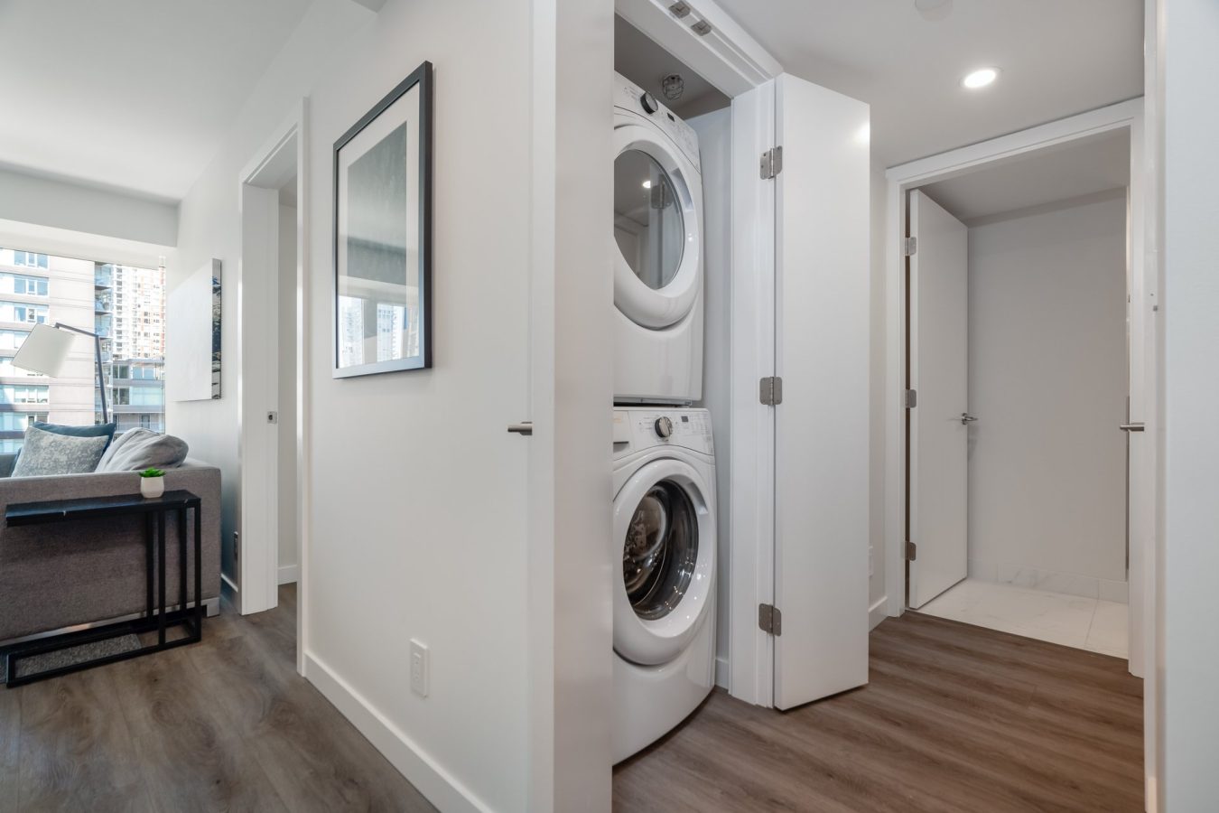 A modern apartment hallway with a stacked washer and dryer in a closet, wood floors, white walls, and a glimpse of a living room with a sofa and large windows in the background.