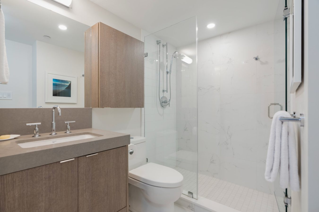 Modern bathroom with a wooden vanity, double sink, wall-mounted cabinet, glass shower enclosure, white toilet, and white tile walls. A towel hangs on the glass shower door and a framed photo is on the wall.