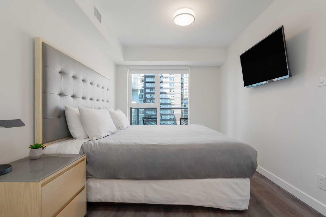 Modern bedroom with a gray upholstered headboard, neatly made bed with white and gray bedding, nightstand with a small plant, large window with a city view, and a wall-mounted flat screen TV.