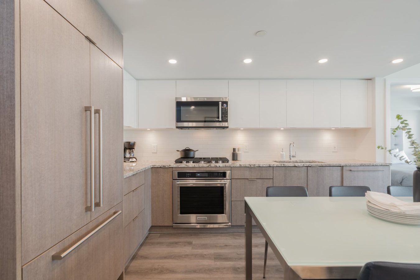 Modern kitchen with light wood cabinets, stainless steel appliances, built-in microwave, gas stove, and a glass dining table with black chairs. Recessed ceiling lights and white backsplash create a bright, clean look.