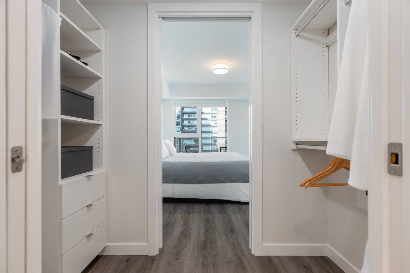 A modern walk-in closet with white shelves, drawers, and hanging space leads to a bright bedroom with a large window, double bed, and cityscape view in the background.
