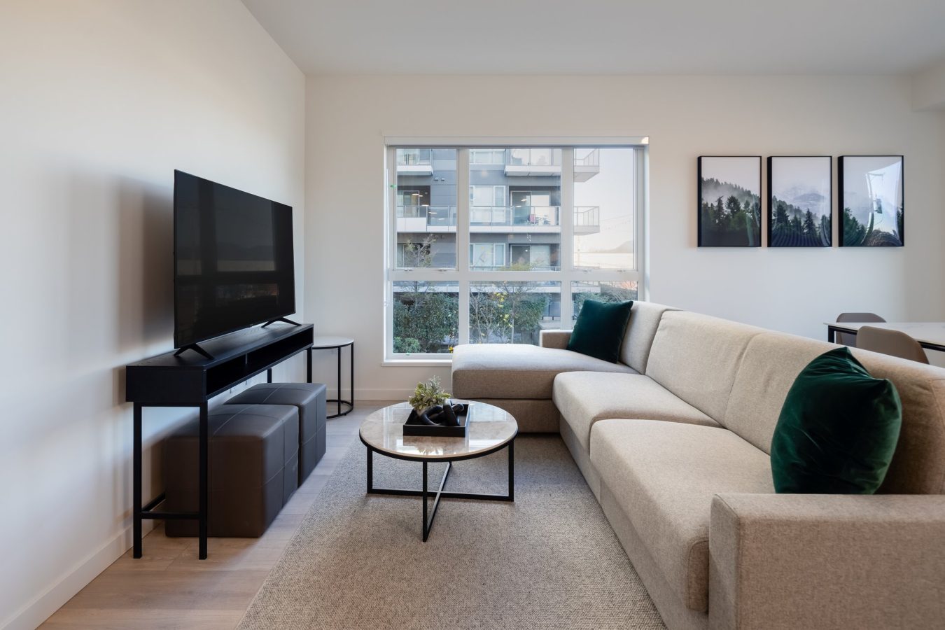 A modern living room with a beige sectional sofa, green pillows, a round coffee table, a TV on a stand, large windows, and framed nature artwork on the wall. An apartment building is visible outside the window.