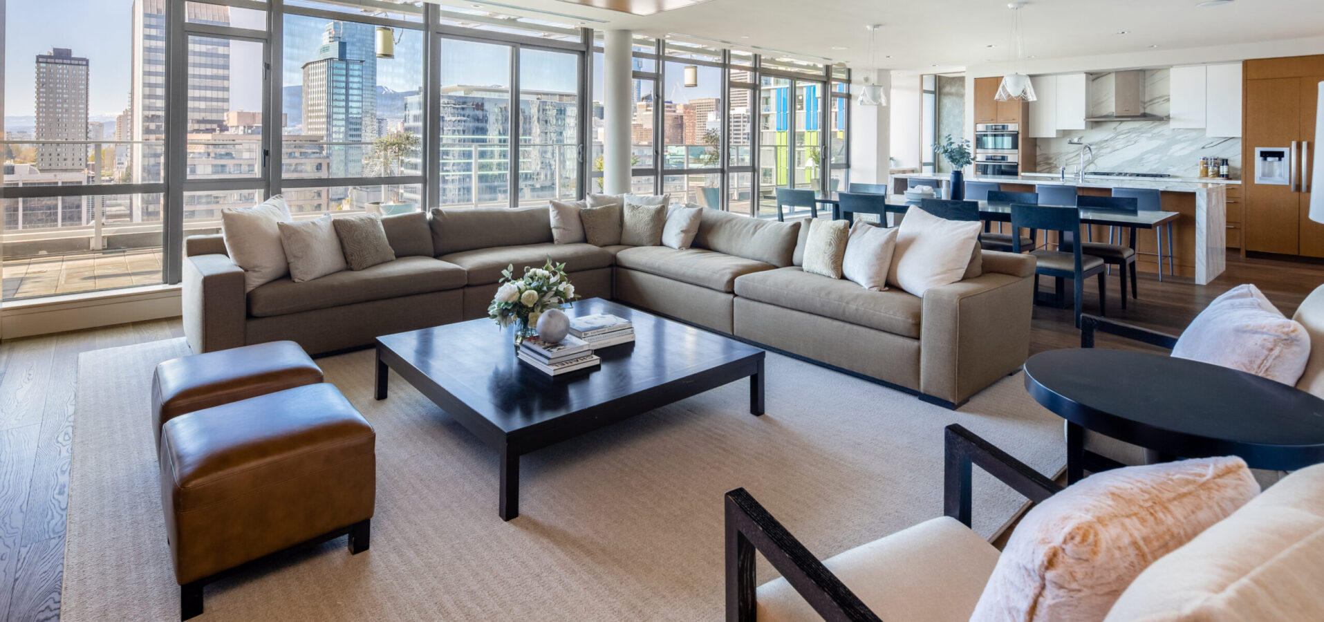 Modern open-concept living room and kitchen with large windows, city views, a beige sectional sofa, brown leather ottomans, a black coffee table, and a dining area with white and blue chairs.