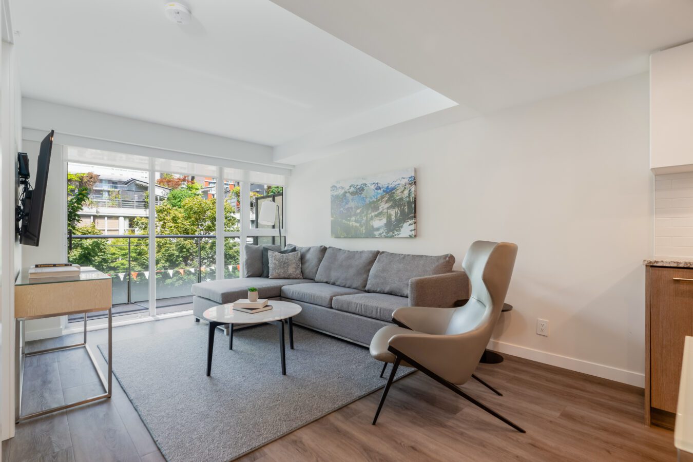 A modern living room with a gray sofa, armchair, coffee table, wall-mounted TV, and large windows letting in natural light. Minimalist decor and wood flooring create a bright, inviting atmosphere.