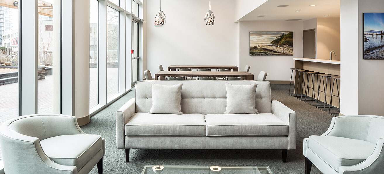 Modern, bright lounge with large windows, a white sofa and armchairs, a glass coffee table, dining table with chairs, bar area with stools, pendant lights, and wall art. Outdoor patio visible through the windows.