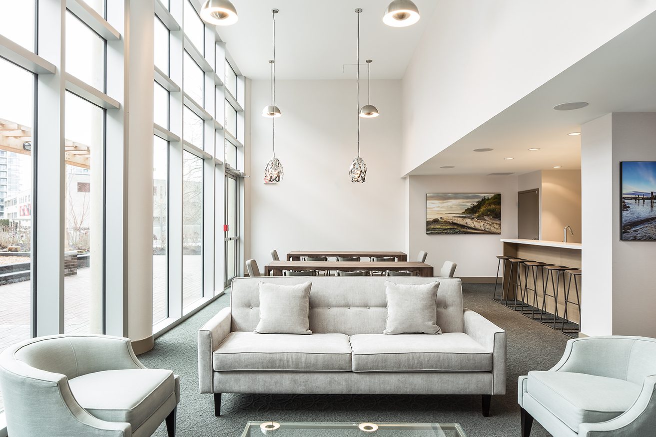 Modern lounge with large windows, a light gray sofa and chairs, a glass coffee table, dining table with chairs, bar counter with stools, pendant lights, and framed landscape photos on white walls. Natural light fills the room.