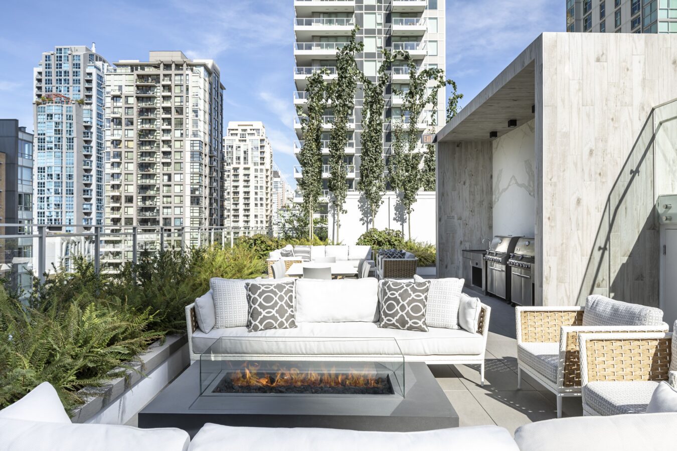 Modern rooftop patio with white sofas, a fire pit, outdoor dining area, built-in grills, and lush plants, surrounded by tall city buildings under a blue sky.