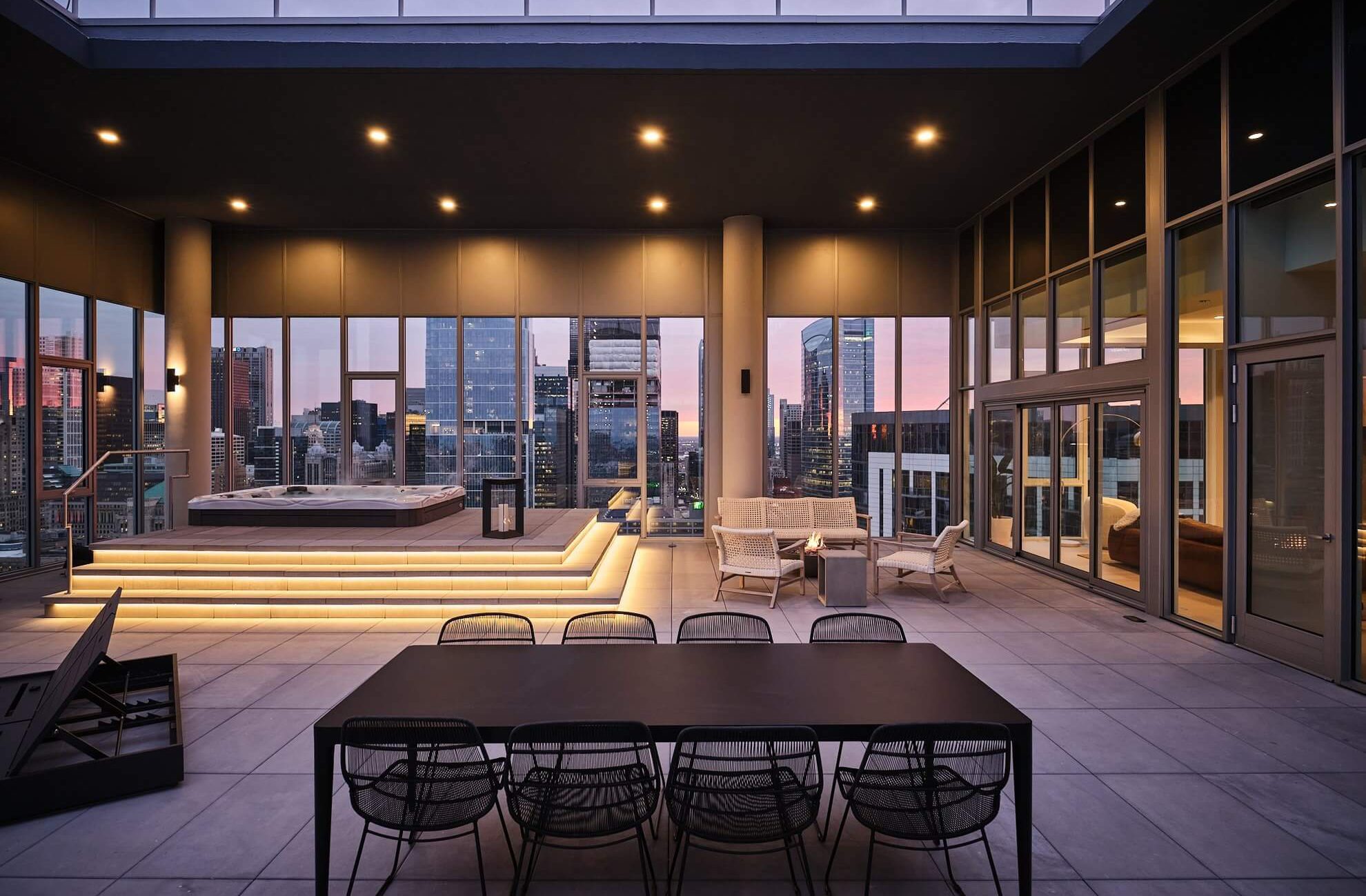 Modern rooftop terrace at dusk with city skyline views, featuring a dining table with chairs, lounge seating, and a raised hot tub with illuminated steps, all surrounded by floor-to-ceiling glass windows.