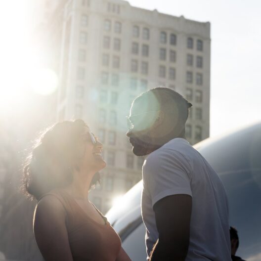 A couple stands close together, smiling at each other in bright sunlight, with a tall building and the reflective surface of Chicago’s Cloud Gate (The Bean) in the background.