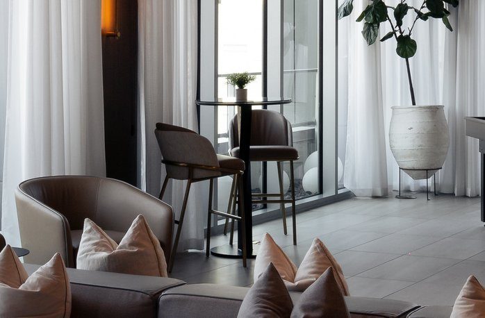 Modern lounge with neutral-toned chairs, a glass table with two barstools, white curtains, a potted plant, and soft lighting, creating a cozy and elegant atmosphere.