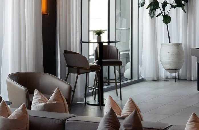 Modern lounge with neutral-toned chairs, a glass table with two barstools, white curtains, a potted plant, and soft lighting, creating a cozy and elegant atmosphere.