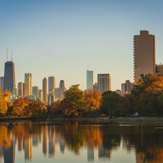 Chicago city skyline with tall buildings set behind trees with autumn foliage, all reflected in the calm waters of a lake under a clear blue sky.