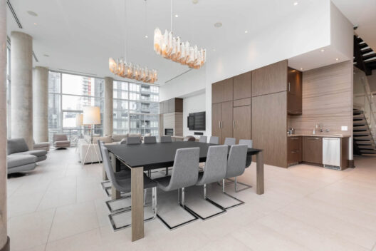 A modern, spacious open-plan living and dining area with floor-to-ceiling windows, a black dining table with gray chairs, wood cabinetry, pendant lighting, and a view of tall city buildings outside.