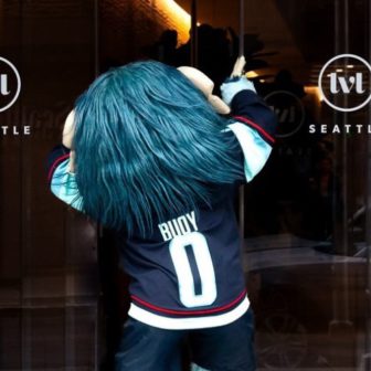 A mascot with long blue hair, wearing a hockey jersey labeled Buoy 0, stands facing a glass door with Seattle and lwl logos. The mascots back is turned to the camera, arms raised.