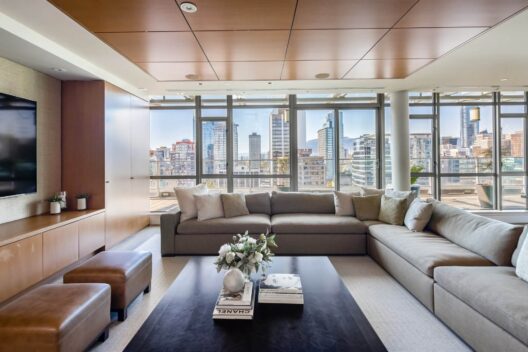 A modern living room with a large gray sectional sofa, two brown ottomans, a black coffee table with books and flowers, and floor-to-ceiling windows overlooking a city skyline.