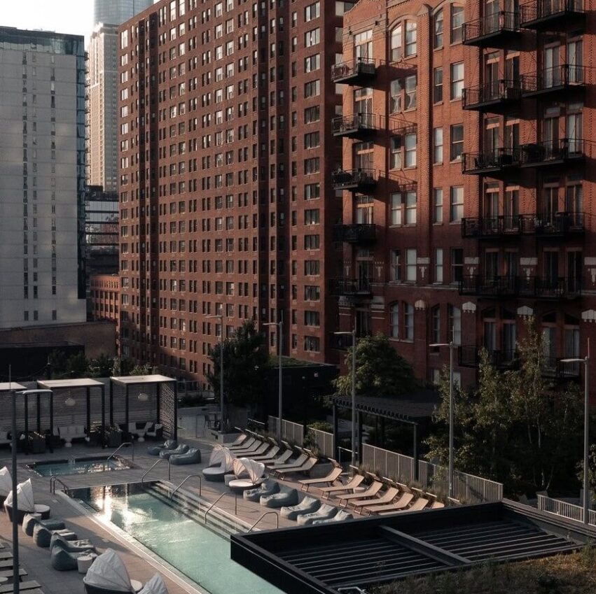 A rooftop pool with lounge chairs and cabanas is surrounded by tall brick and glass apartment buildings in a city setting, lit by soft evening sunlight.