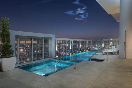 A modern rooftop pool at night overlooks a city skyline with sparkling lights. The pool is surrounded by lounge chairs, potted plants, and glass barriers; a swirling cloud pattern is visible in the dark sky above.