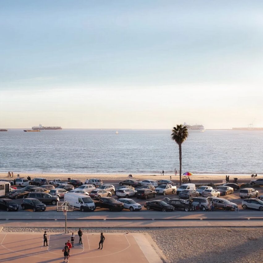 A beachside parking lot is crowded with cars near a sandy shore. People play basketball in the foreground, while ships are visible on the calm ocean under a clear sky. A single palm tree stands by the beach.