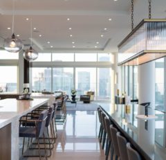 Modern, spacious kitchen and dining area with a glossy island, high-backed stools, contemporary pendant lights, and large windows offering a city skyline view. A bright living area with armchairs is visible in the background.