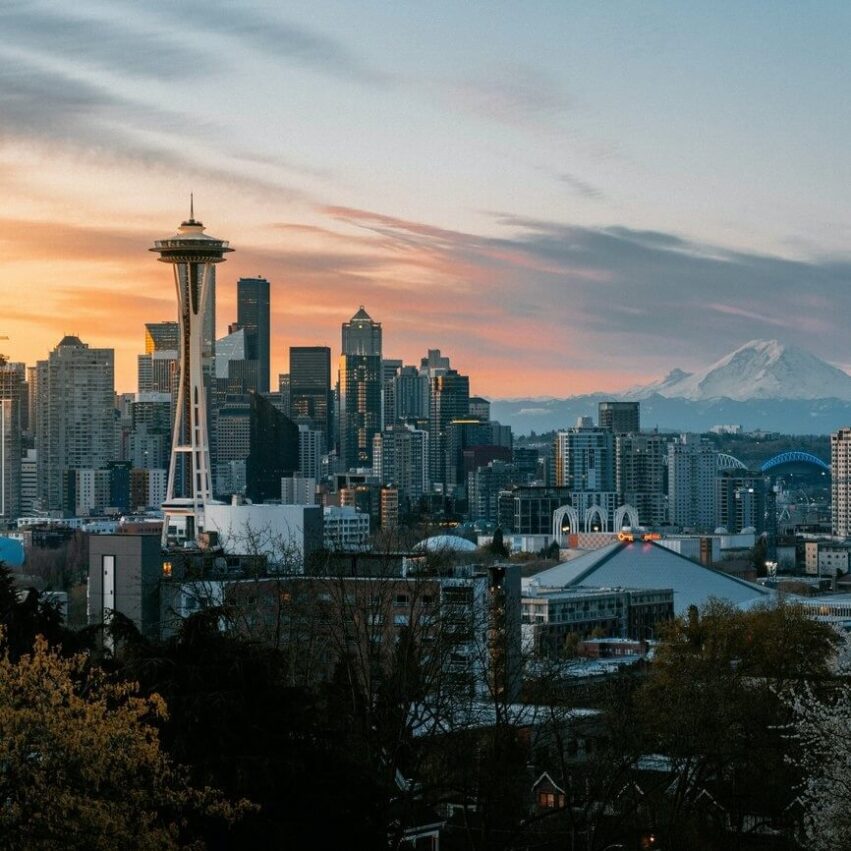 Seattle skyline at sunset, featuring the Space Needle in the foreground, downtown skyscrapers, and Mount Rainier in the distance under a partly cloudy sky. Trees and buildings fill the lower part of the image.