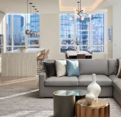 Modern, sunlit open-plan living space with a light gray sofa, decorative vases on round coffee tables, a dining area, and a kitchen island with bar stools. Large windows reveal a city skyline view.