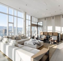 A modern, spacious living room with floor-to-ceiling windows, white sofas, a large ottoman, a light rug, a dining area with high-back chairs, and panoramic city views with mountains in the background.