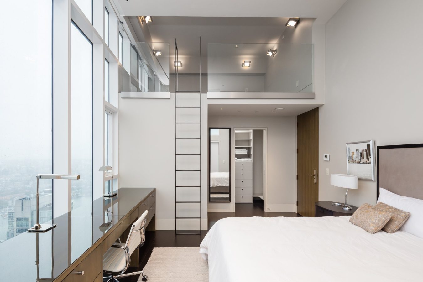 Modern bedroom with a large window, desk and chair along one wall, a bed with pillows, and a tall ladder leading to a glass-railed loft area above a walk-in closet. Neutral colors and minimalist decor.