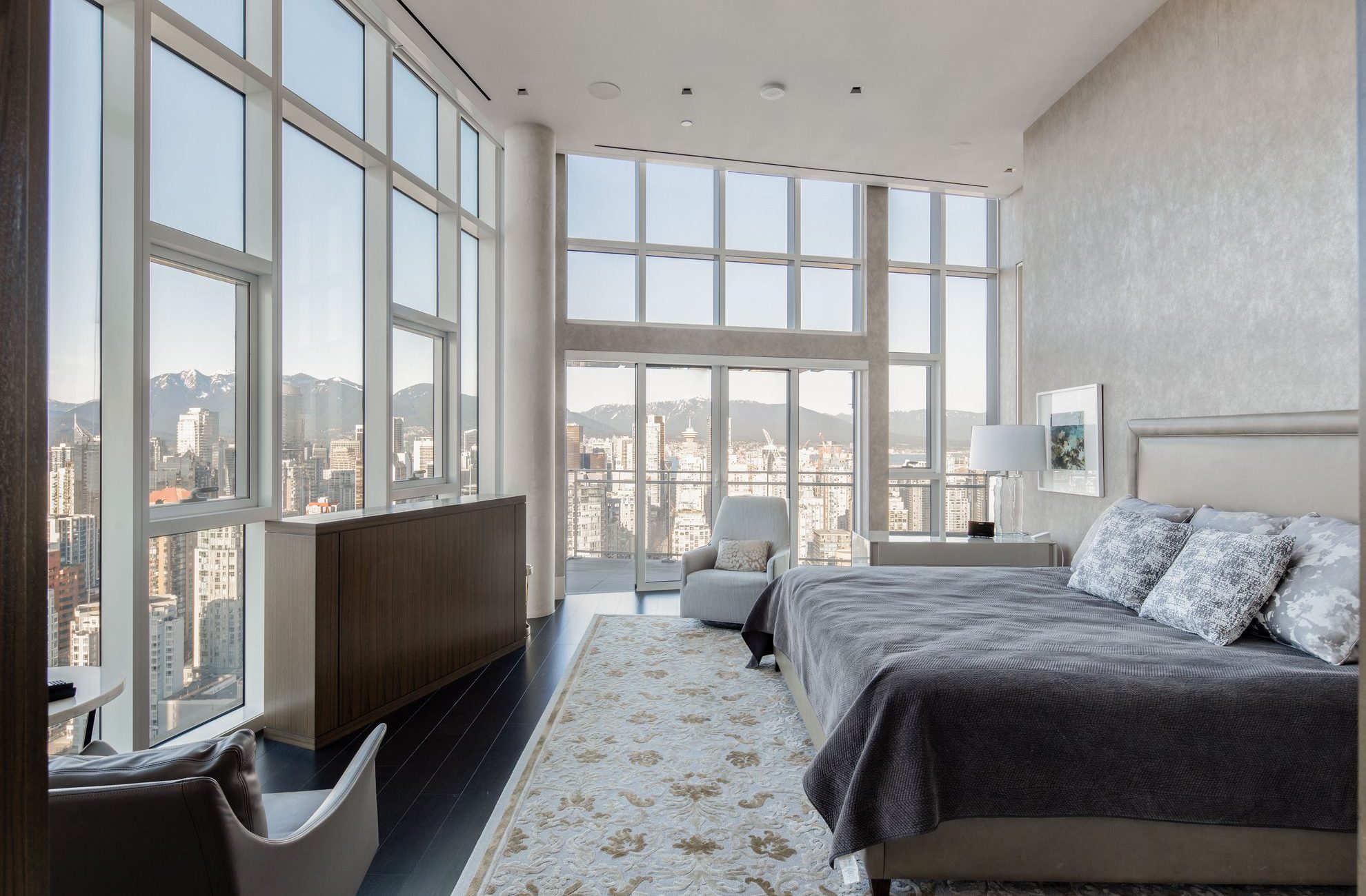 A modern bedroom with large floor-to-ceiling windows offers a city skyline and mountain view, with a bed, armchair, light rug, and neutral decor in a bright, airy space.