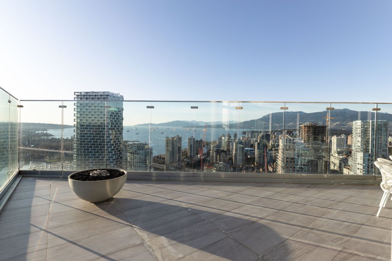 Modern rooftop terrace with glass railing overlooking a city skyline, ocean, and distant mountains on a clear day. A round planter sits on the tiled floor; some chairs are partially visible on the right.