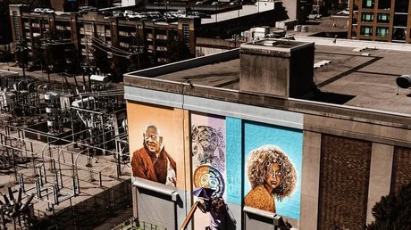 Aerial view of a city building with three large, colorful murals depicting diverse faces on its exterior. Nearby, power lines and industrial structures are visible.