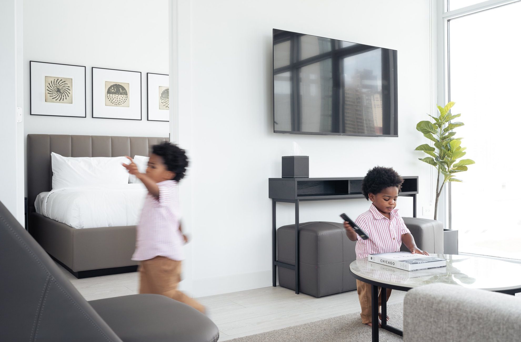 Two young children are in a modern, bright living room. One child sits at a glass coffee table with a TV remote, while the other walks towards a bed in an adjacent room. The space is decorated with art, plants, and a large wall-mounted TV.