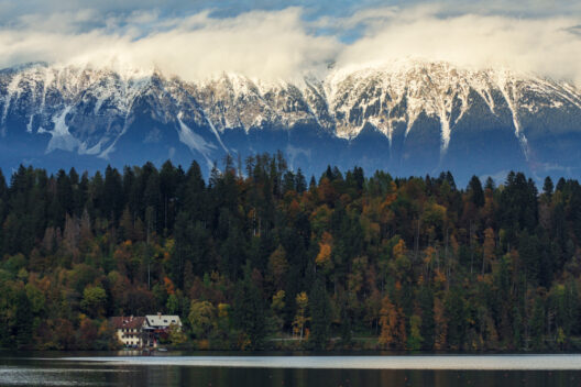 A peaceful lakeside scene with a house nestled among dense autumn trees. Behind the forest, tall mountains rise, topped with snow and partially covered by clouds under a dramatic sky.