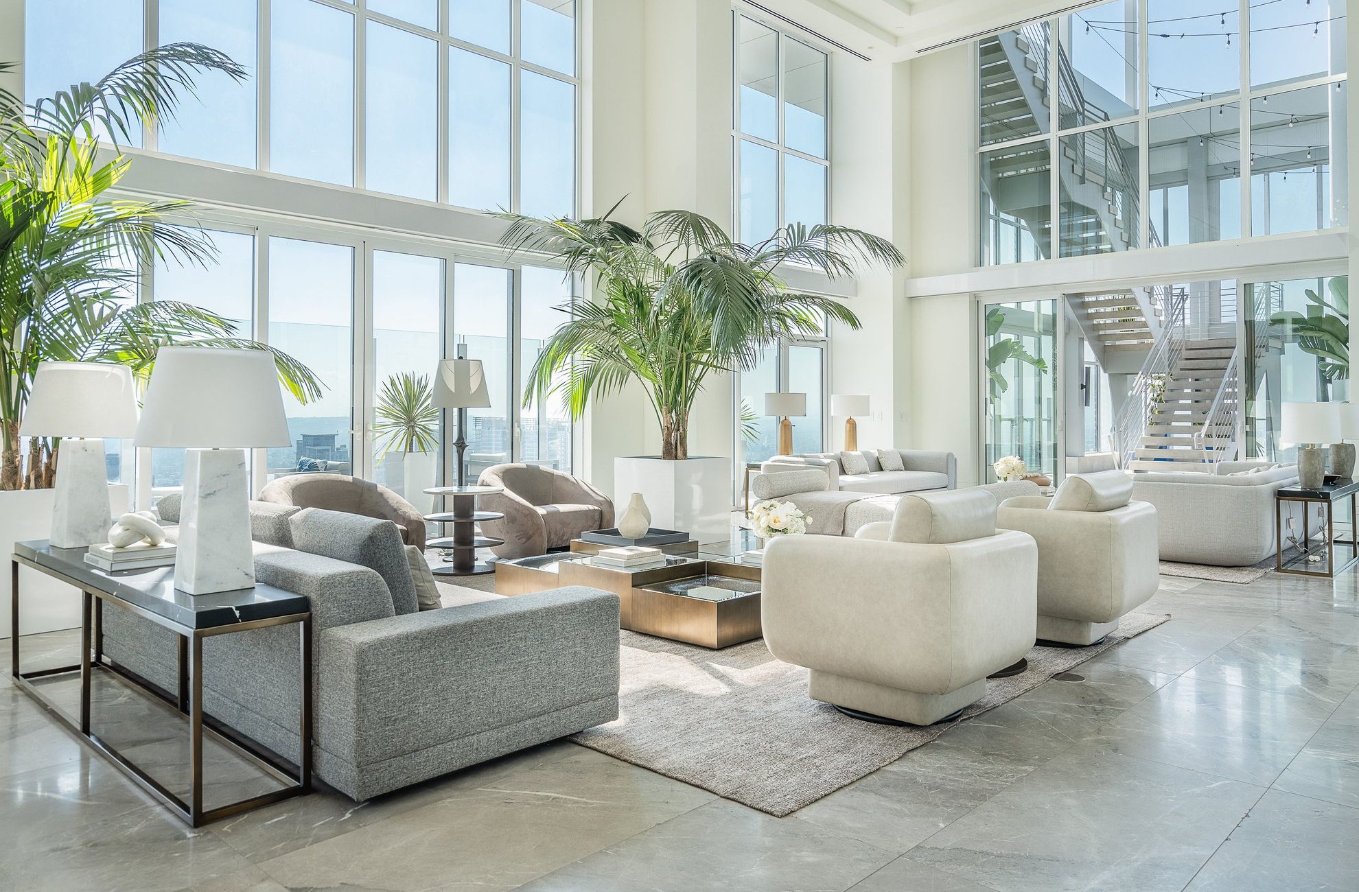 A bright, spacious modern living room with large windows, neutral-toned sofas and armchairs, glass coffee tables, potted palm plants, and a staircase leading to an upper level. Natural sunlight fills the space.