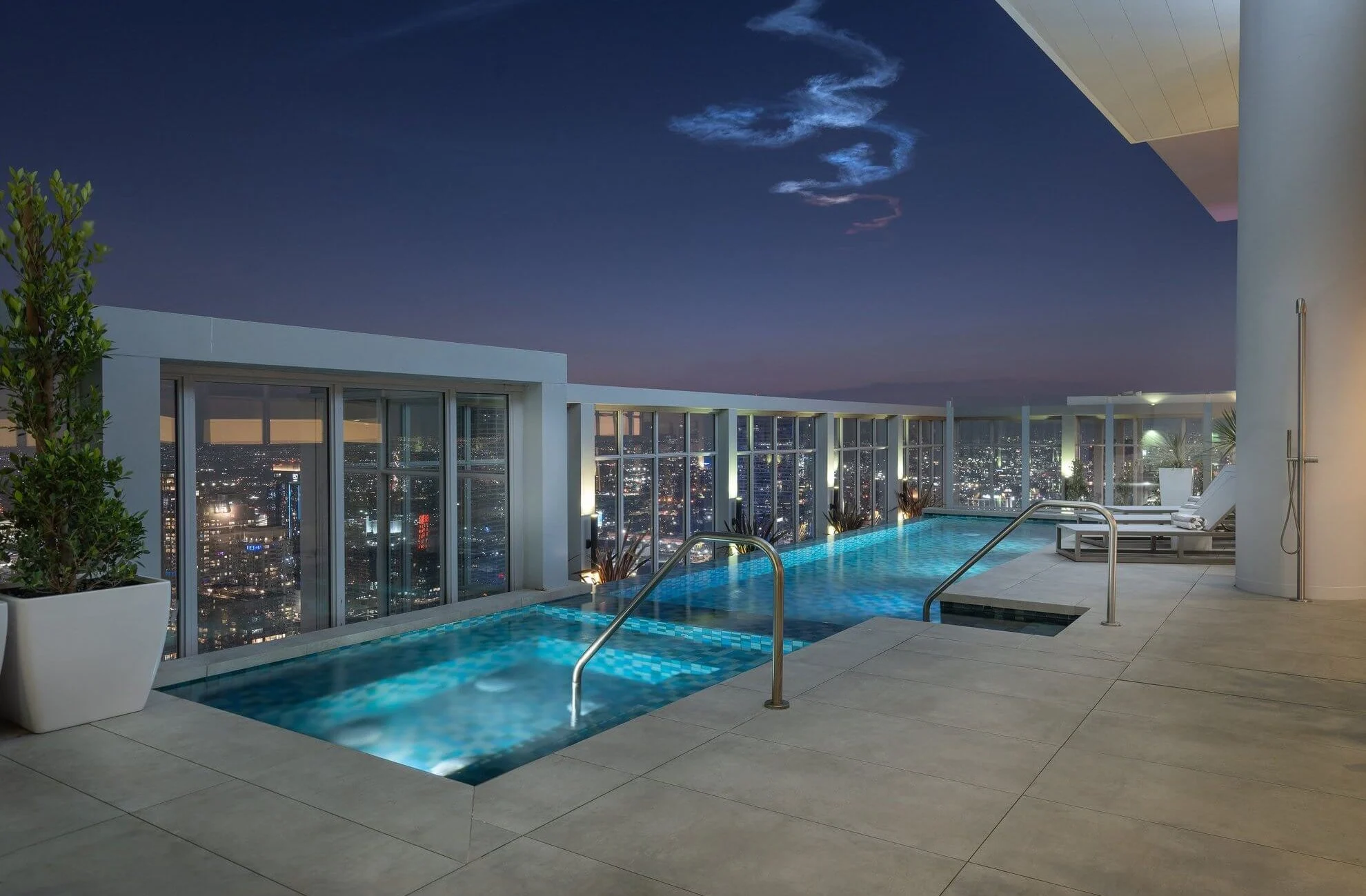 A modern rooftop infinity pool overlooking a city skyline at night, with glass railings, lounge chairs, and potted plants, all illuminated by soft lighting under a twilight sky.