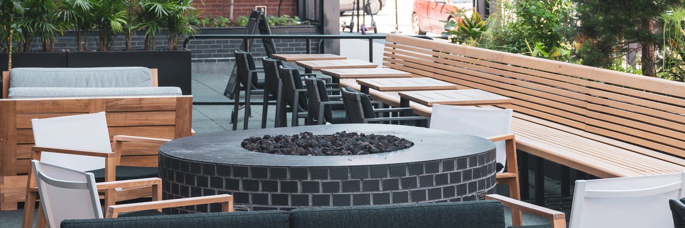 Outdoor patio with modern seating, a large round black brick fire pit in the center, wooden benches along the side, tables with chairs, and plants in the background.