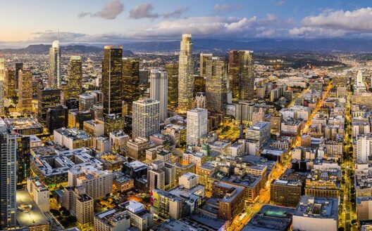 Aerial view of downtown Los Angeles at dusk, showing tall skyscrapers, city lights, and surrounding urban buildings under a partly cloudy sky.