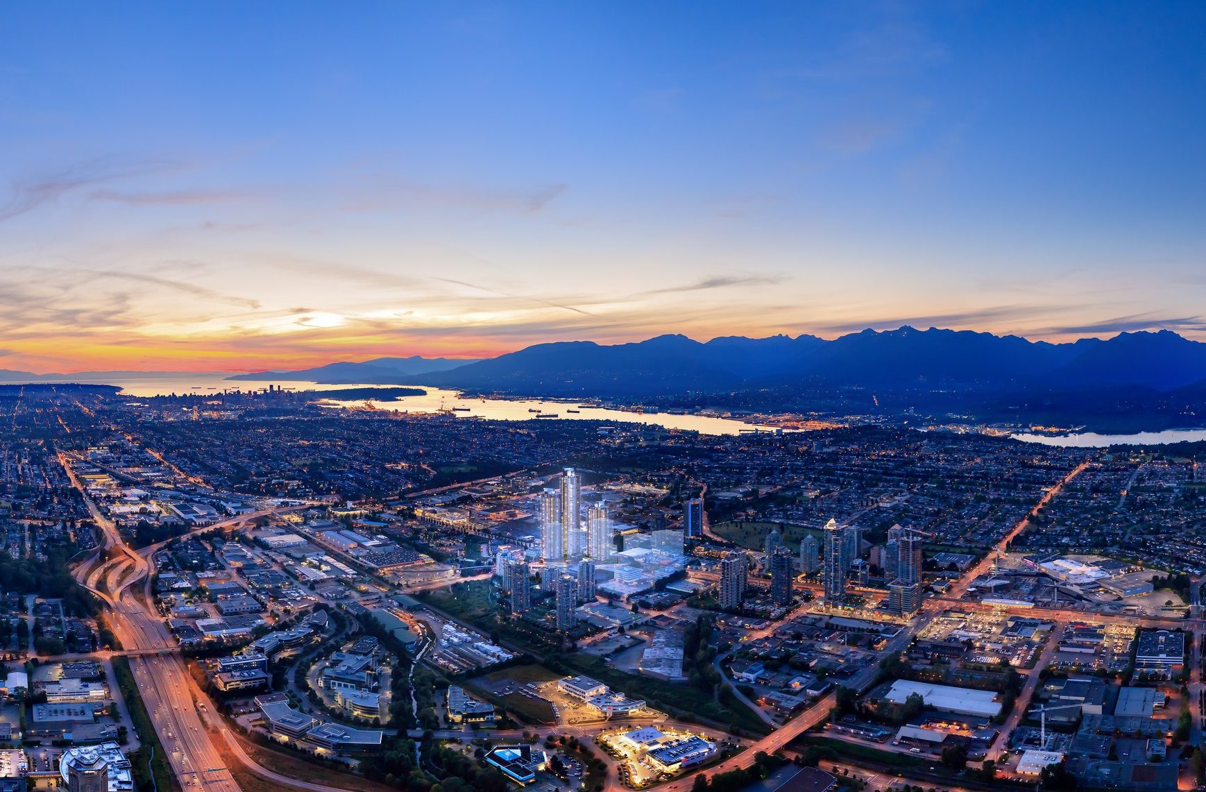 Aerial view of a city at sunset, with glowing skies, distant mountains, a river, and illuminated buildings and roads below. Urban and natural elements blend under a tranquil twilight.