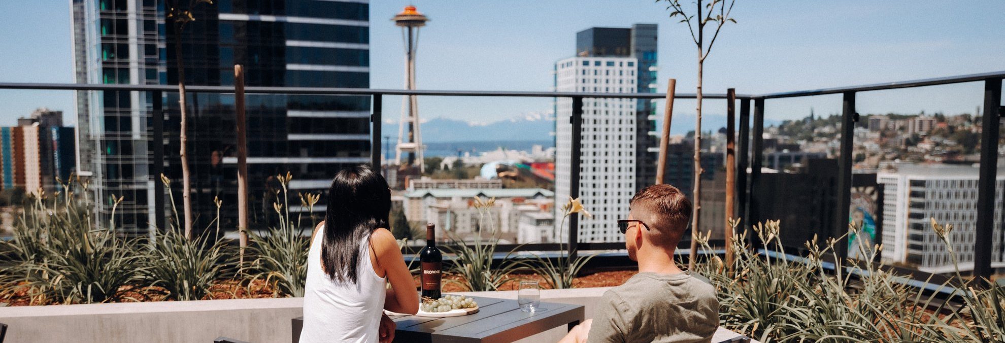 Two people sit at a rooftop table overlooking downtown Seattle, with the Space Needle and surrounding skyscrapers visible in the background under a clear sky.