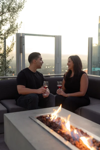 A man and woman sit on a rooftop patio by a fire pit, smiling at each other and holding glasses of wine at sunset. The cityscape is visible in the background through glass barriers.