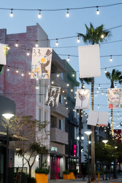 A city street scene at dusk with hanging string lights and illustrated banners, palm trees, modern buildings, and the pink T-Mobile sign visible above a storefront.