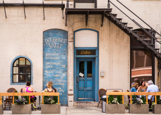 Outdoor seating in front of a bakery with a blue door, menu painted on the wall, potted plants, and people sitting at tables under a fire escape. The sign reads “Doughnut Vault” and lists various doughnut flavors.