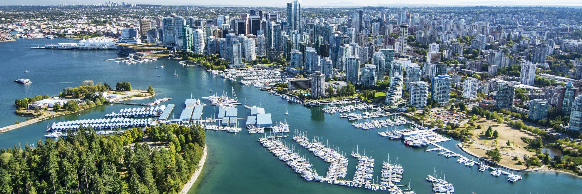 Aerial view of a city skyline with numerous high-rise buildings, dense urban areas, a large marina filled with boats, and a green park area by the waterfront.