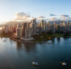 A panoramic view of Vancouver’s downtown skyline at sunset, with high-rise buildings, marina boats, calm water, and mountains in the background under a partly cloudy sky.