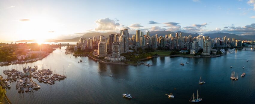 A panoramic view of Vancouver’s downtown skyline at sunset, with high-rise buildings, marina boats, calm water, and mountains in the background under a partly cloudy sky.
