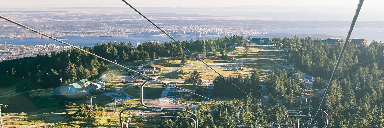 A scenic view from a ski lift overlooking a green mountainside, city buildings, water with ships, and distant land on the horizon under a clear sky.